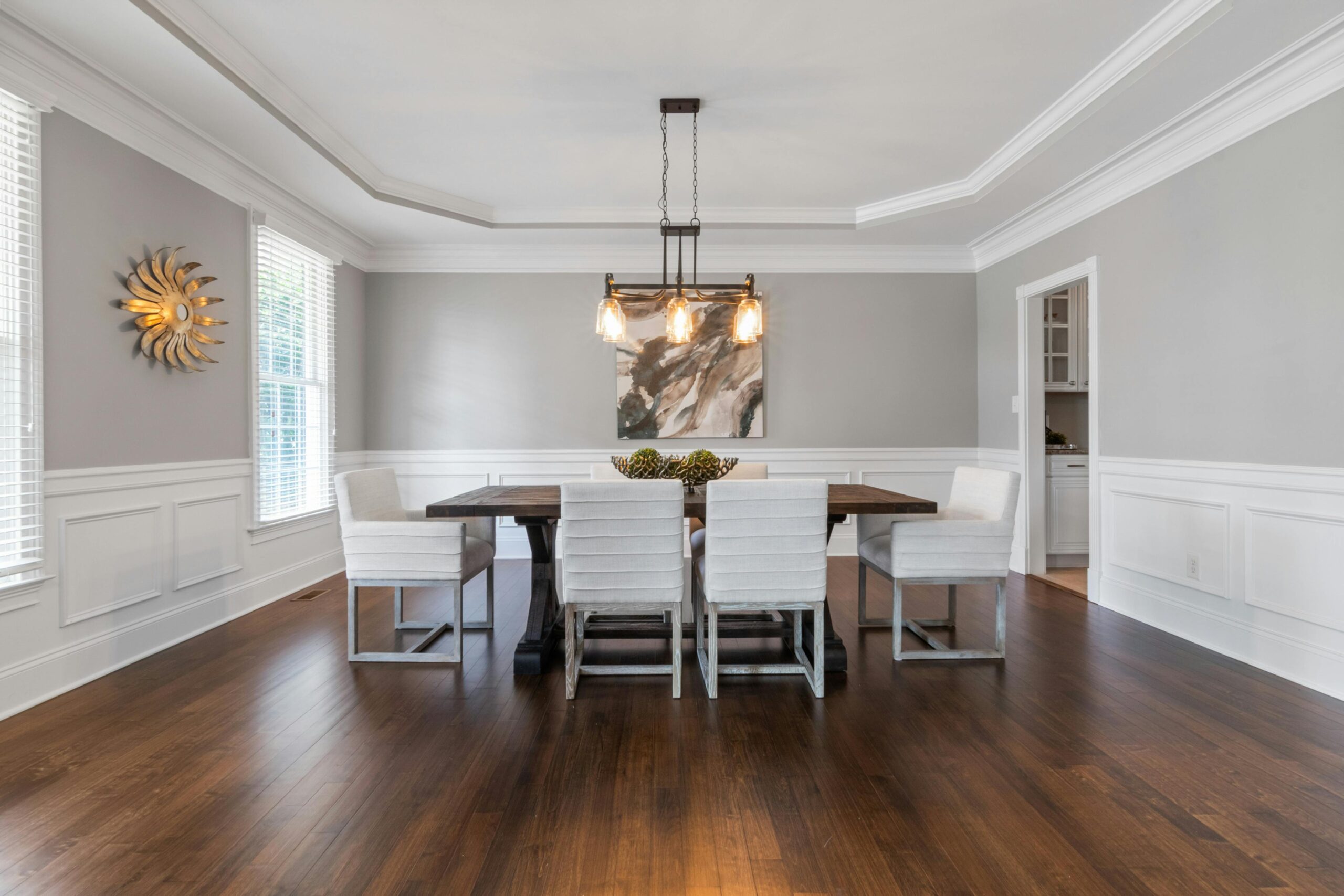 Modern dining room featuring rich dark hardwood flooring with wide planks, white wainscoting, gray walls, and a rectangular wood table with upholstered chairs under a contemporary pendant light.