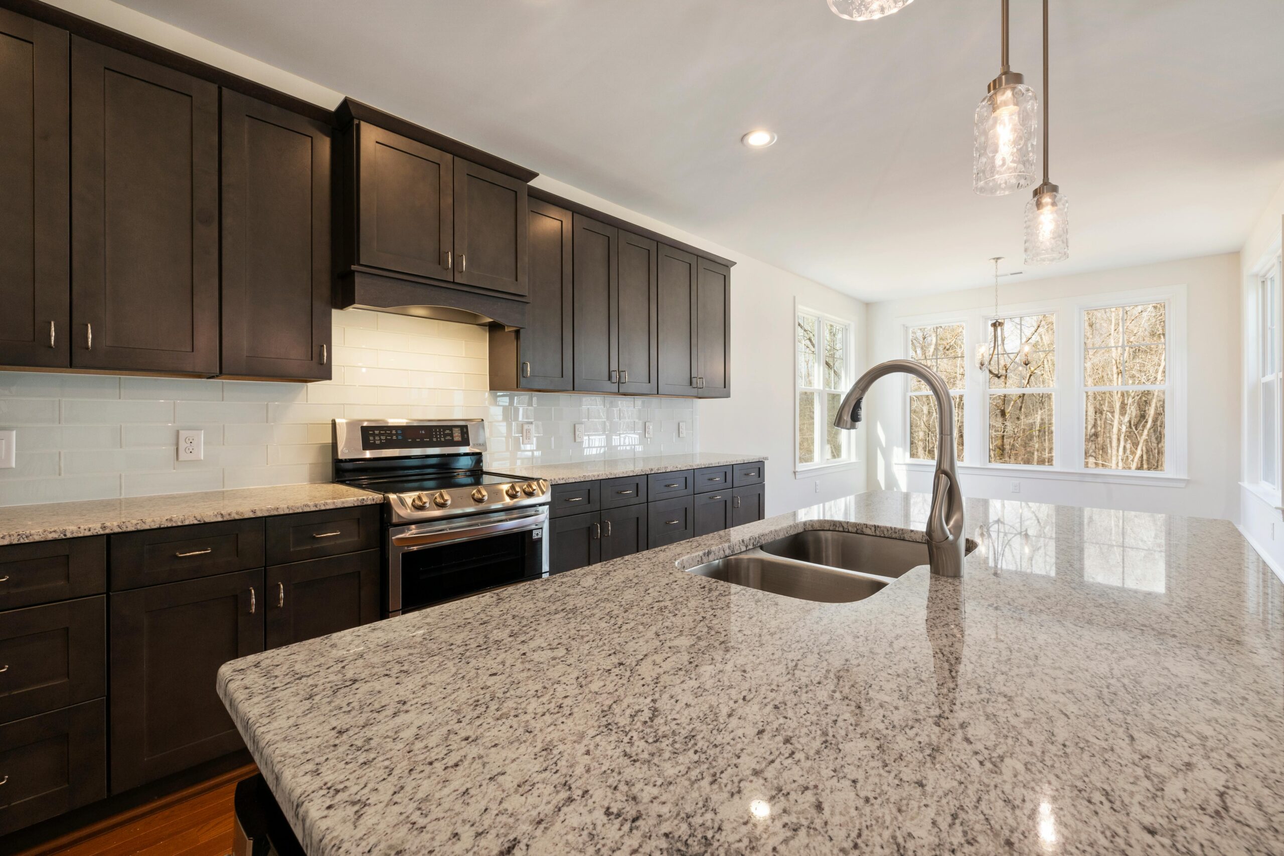 Modern kitchen featuring glossy white subway tile backsplash, dark espresso cabinets, granite countertops, and stainless steel appliances with natural light from large windows.